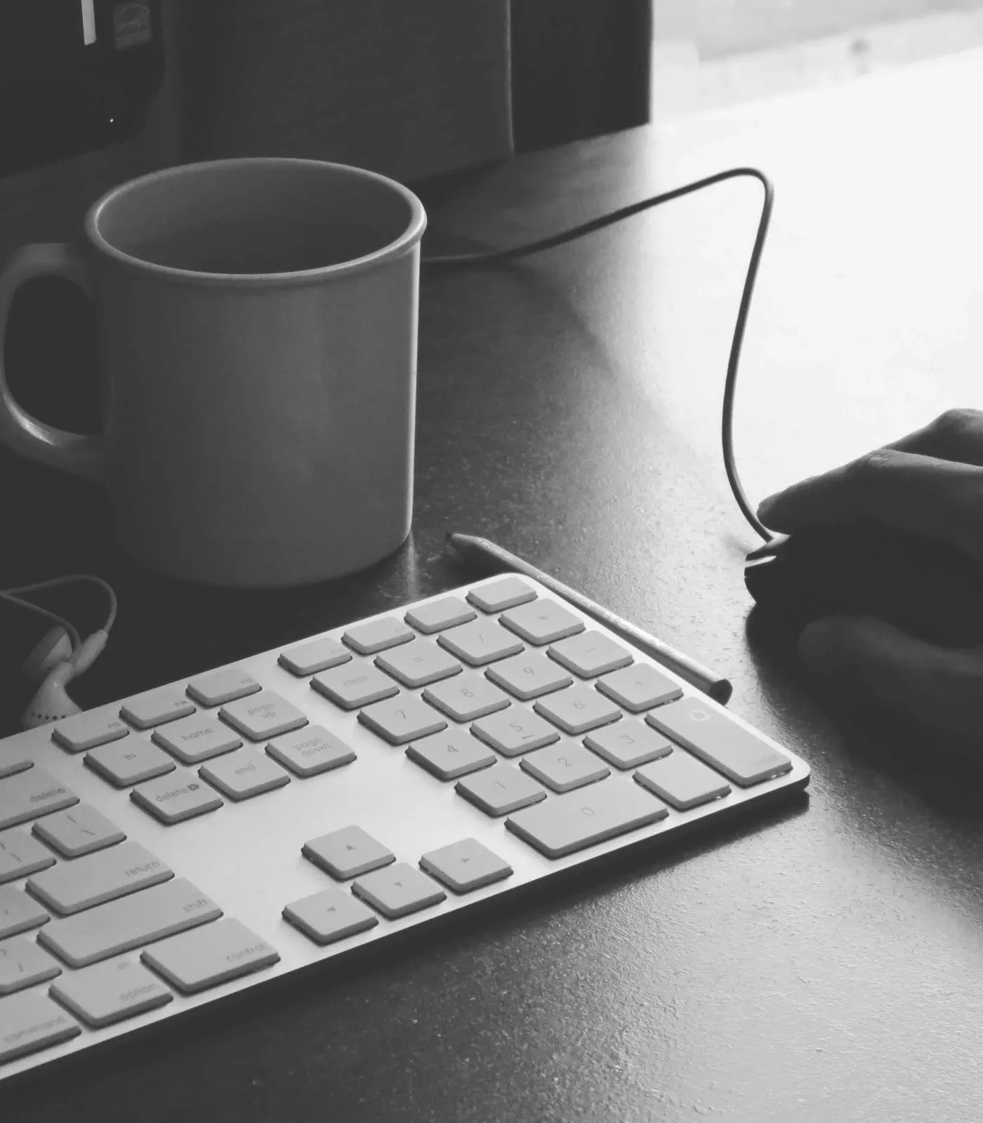 Black and white photo of a hand on a mouse beside a keyboard and coffee mug